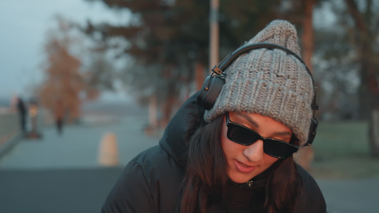 Primo piano di una donna che balla con le cuffie, berretto di lana e occhiali da sole che incorniciano il viso sorridente. Indossa un cappotto invernale, con uno skyline sul lungomare sullo sfondo. I movimenti della testa si sincronizzano con la musica, creando un'atmosfera serale urbana accogliente e spontanea.
