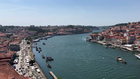 Panoramic-shot-over-city-of-Oporto-with-Douro-River,-old-town-buildings-and-rabelo-boats-during-sunny-day-with-blue-sky---view-from-top-of-Ponte-Dom-Luís-I,-Portugal