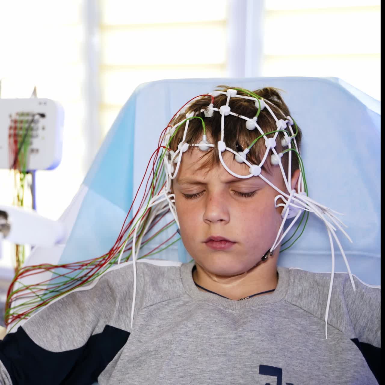 EEG examination for a teenage boy. Little patient wearing sensors frowns as the light flashes. Doctor sitting at backdrop in blur
