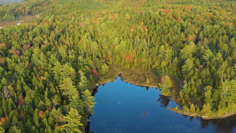 aerial drone orbiting around a bright blue lake with colorful autumn trees surrounding the water as summer ends and the seasons change to fall in maine