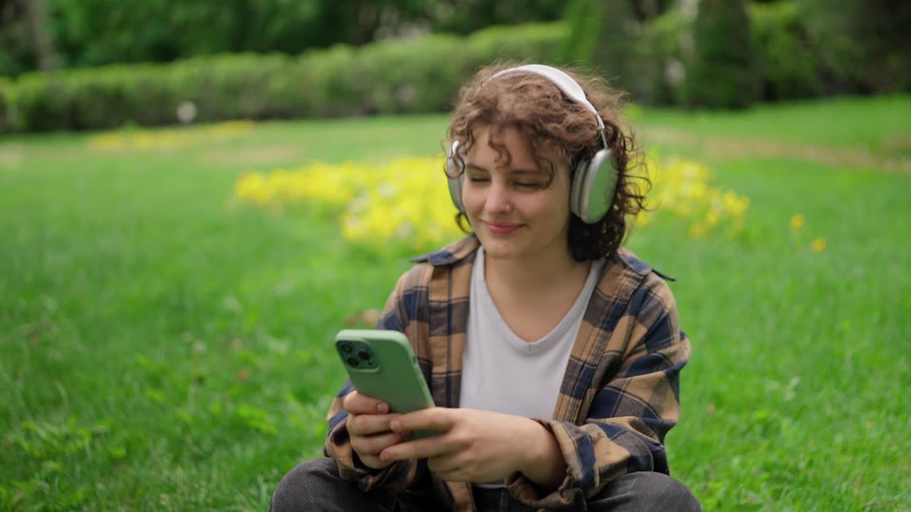 feliz chica morena con cabello rizado se sienta en la hierba verde escuchando música en auriculares en un parque en verano
