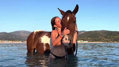 young red-haired girl in bikini hugs and kisses and cuddles horse bathing in sea water in summer season