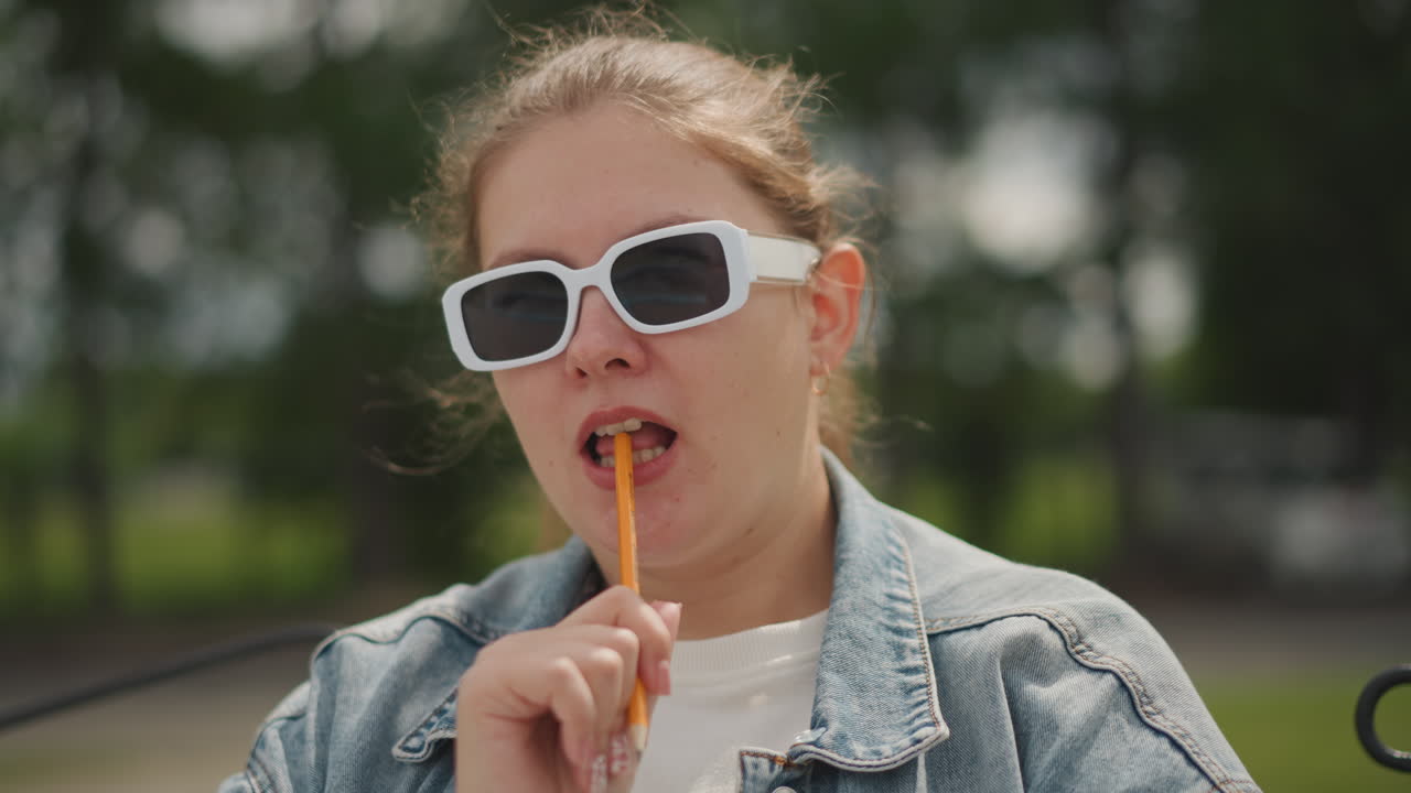 Caucasian girl in sunglasses chewing straw, denim jacket on park bench, pensive student sketching ideas, soft sunlight, blurred trees background, candid closeup capturing contemplative summer mood