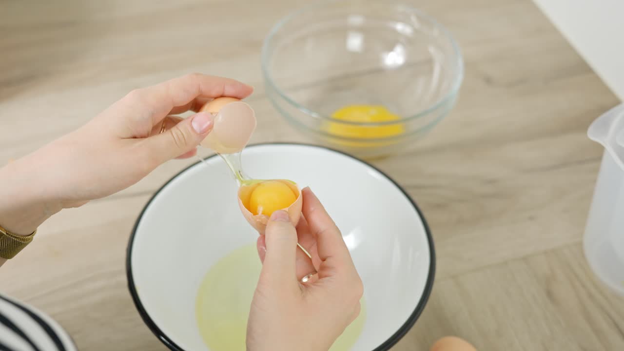 Separating egg yolk by hands into white bowl on wooden kitchen table during cooking, food preparation in process for baking making desert or homemade recipe