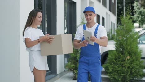 una mujer hermosa conoce a un repartidor que le da una caja de paquetes al lado de su casa, ella firma una firma electrónica en una tableta. concepto de mensajero, entrega a domicilio, compras en línea