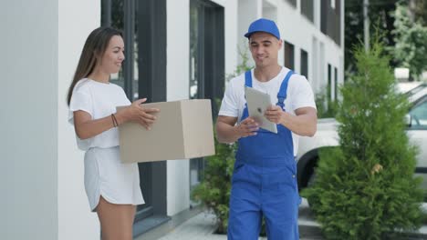 beautiful woman meets a delivery man who gives her a parcel box beside her home, she signs electronic signature on tablet. concept of courier, home delivery, online shopping