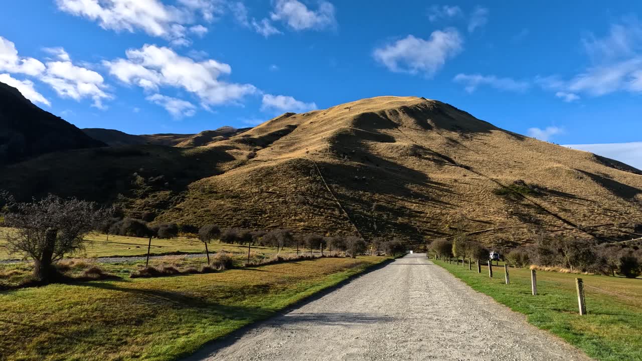 Vehicle moves down rural gravel road, passing fields and trees, under bright daylight, steady camera