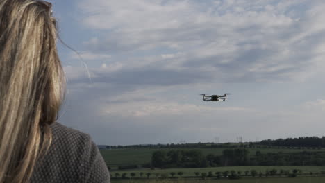 Back-View-of-Young-Woman-Controlling-Drone-Flying-Under-Sky-on-Sunny-Day,-Slow-Motion