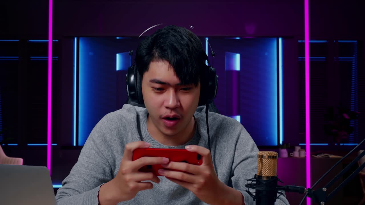 primer plano de un feliz jugador asiático con auriculares y computadora celebrando la victoria en el juego de teléfonos móviles en una habitación iluminada por luces de neón en retro