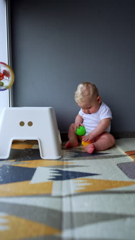 adorable plump caucasian baby in white bodysuit stands at the chair hitting it with a ball. cute kid drops on the floor playing with toys. vertical video.