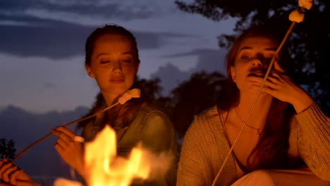 two young caucasian girls sit in the evening by the fire in nature, eating marshmelow, looking at an open fire, thinking