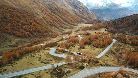 aerial view of a winding mountain road surrounded by autumn-colored trees and rugged terrain, with snow-capped peaks in the background, creating a picturesque and serene landscape
