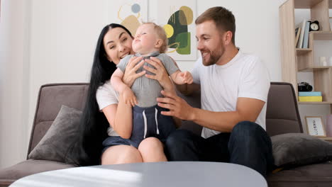 Padres-Amorosos-Sentados-En-Un-Sofá-En-La-Sala-De-Estar-Jugando-Con-Su-Adorable-Niño-Mientras-Miran-La-Cámara-Durante-Una-Videollamada-En-Casa