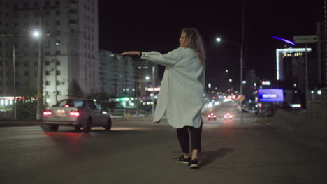 woman in white coat standing by roadside at night raising hand to get taxi as silver car drives past without stopping on urban street surrounded by city lights and high-rise buildings