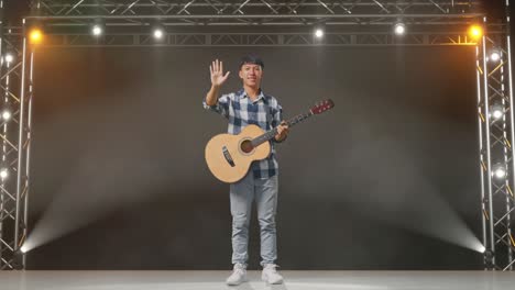 cuerpo lleno de adolescente asiático con la mano agitando la guitarra y sonriendo a la cámara en el escenario