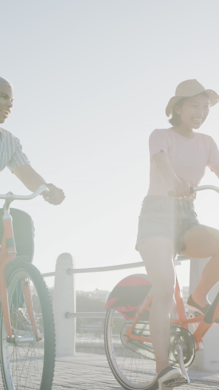 Vertical video of happy biracial couple hriding bikes on promenade, in slow motion
