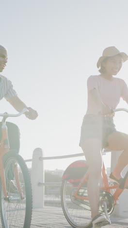 vertical video of happy biracial couple hriding bikes on promenade, in slow motion