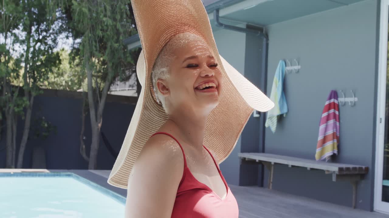 retrato de una feliz mujer biracial con sombrero de sol en la piscina en cámara lenta