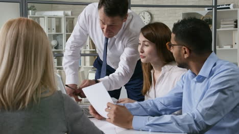 senior business mentor showing something on laptop and talking to young diverse coworkers at meeting table in the office; business lady giving an advice