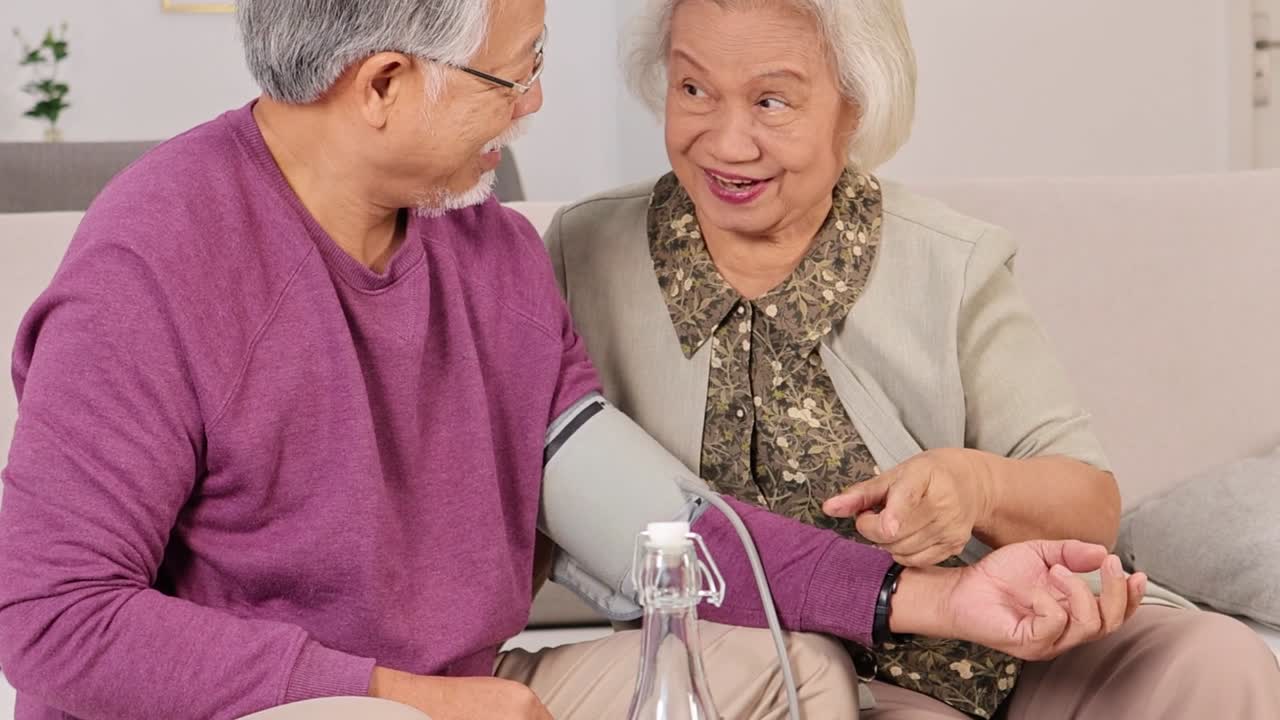 Un uomo e una donna anziani sorridono mentre monitorano la pressione arteriosa su un divano in salotto.