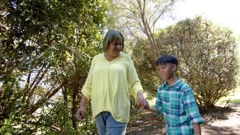 happy biracial grandmother and grandson walking in sunny garden, slow motion