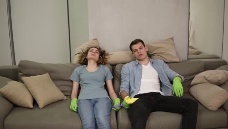 exhausted young caucasian couple falling down on a couch after cleaning, feeling tired after cleaning, swiping sweat from foreheads. exhaling. wearing green rubber gloves. slow motion