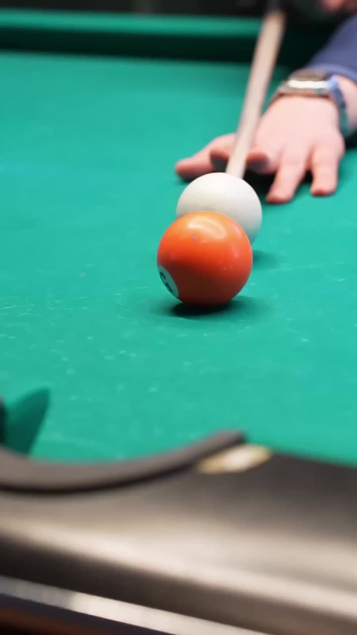 Primo piano verticale di una stecca da biliardo che colpisce la palla bianca mentre una palla arancione rotola nella buca d'angolo su un tavolo di feltro verde, catturando un tiro riuscito al chiuso