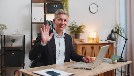 happy caucasian businessman employee waves hand hi gesture sitting with laptop at home office desk