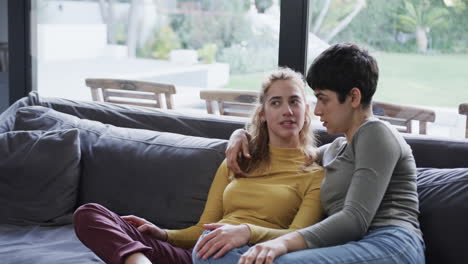 feliz pareja de lesbianas caucásicas sentadas en el sofá, abrazándose y hablando en la sala de estar soleada