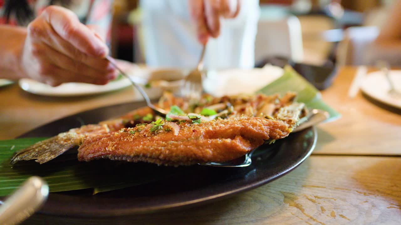 Hand pours spicy herb sauce over crispy fried fish in bright Bangkok restaurant, shallow focus