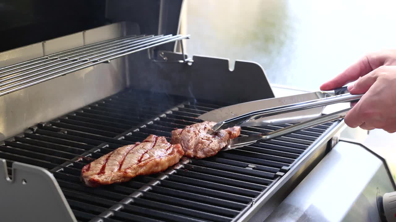 A person grills a beef steak on a barbecue, using tongs to flip it. Bright lighting enhances the cooking process