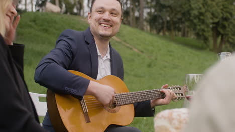 middle aged man playing a guitar sitting at table with his friends during an outdoor party in the park