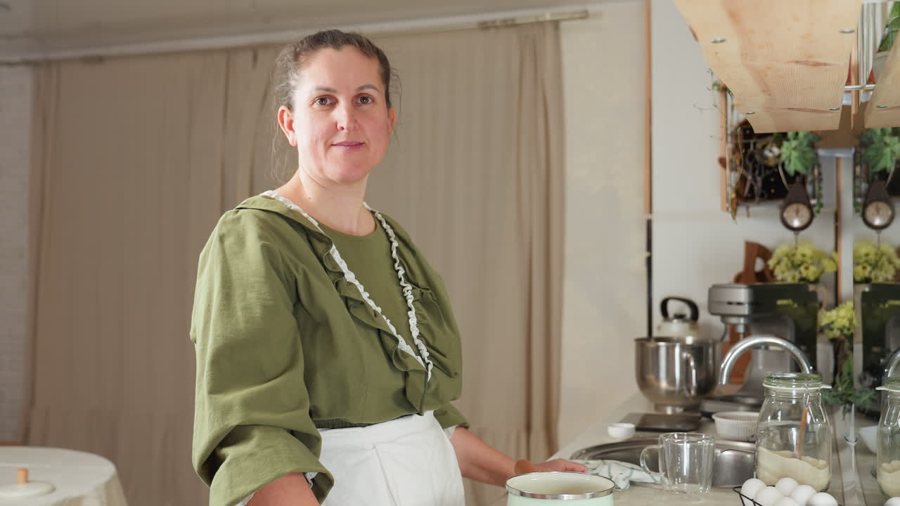 Cook in green dress and white apron pauses in kitchen work to smile gently at camera while standing beside countertop with pot, eggs, and flour jar, in warmly lit rustic kitchen