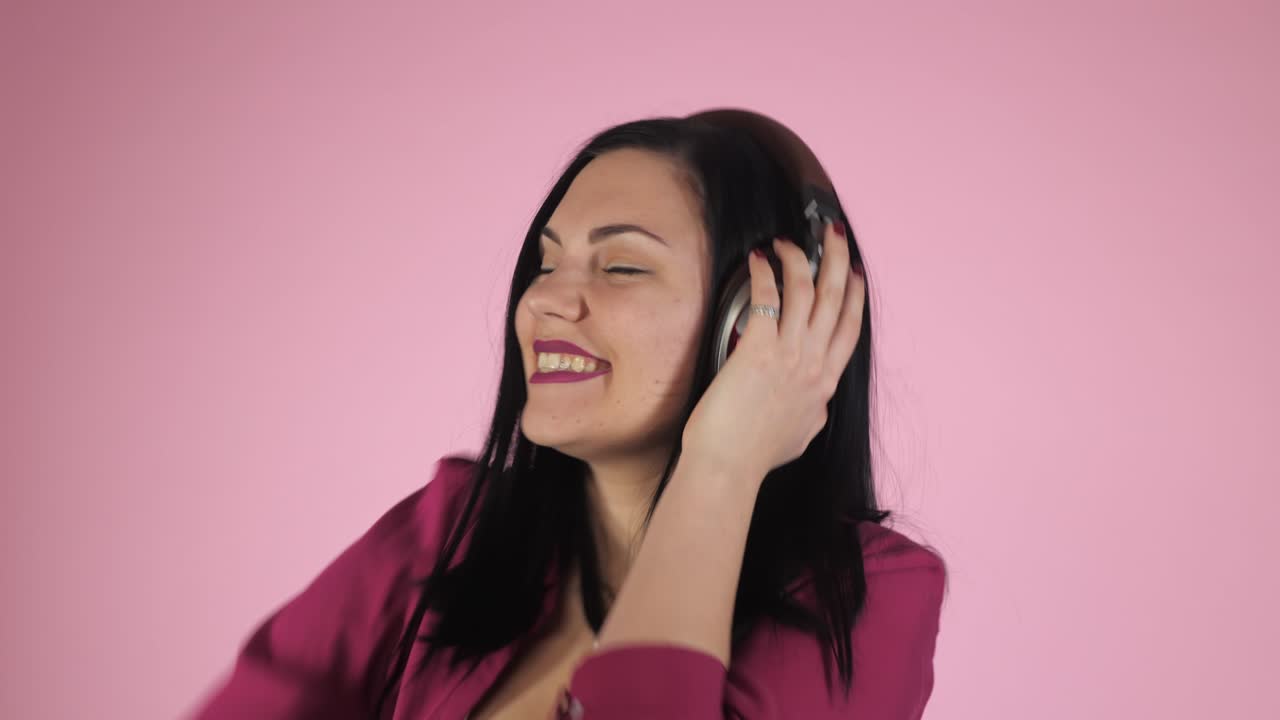 Bella giovane donna con le cuffie che ascolta musica e canta a occhi chiusi, in piedi su uno sfondo rosa, in camicia. 4k.