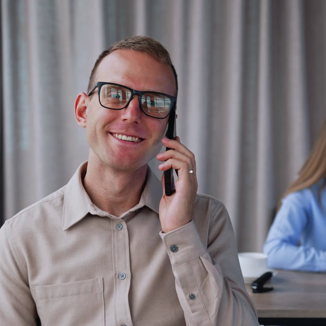 Having a phone call in office. Man speaks on his phone smiling. Close up. Lady at backdrop uses phone and laptop at once
