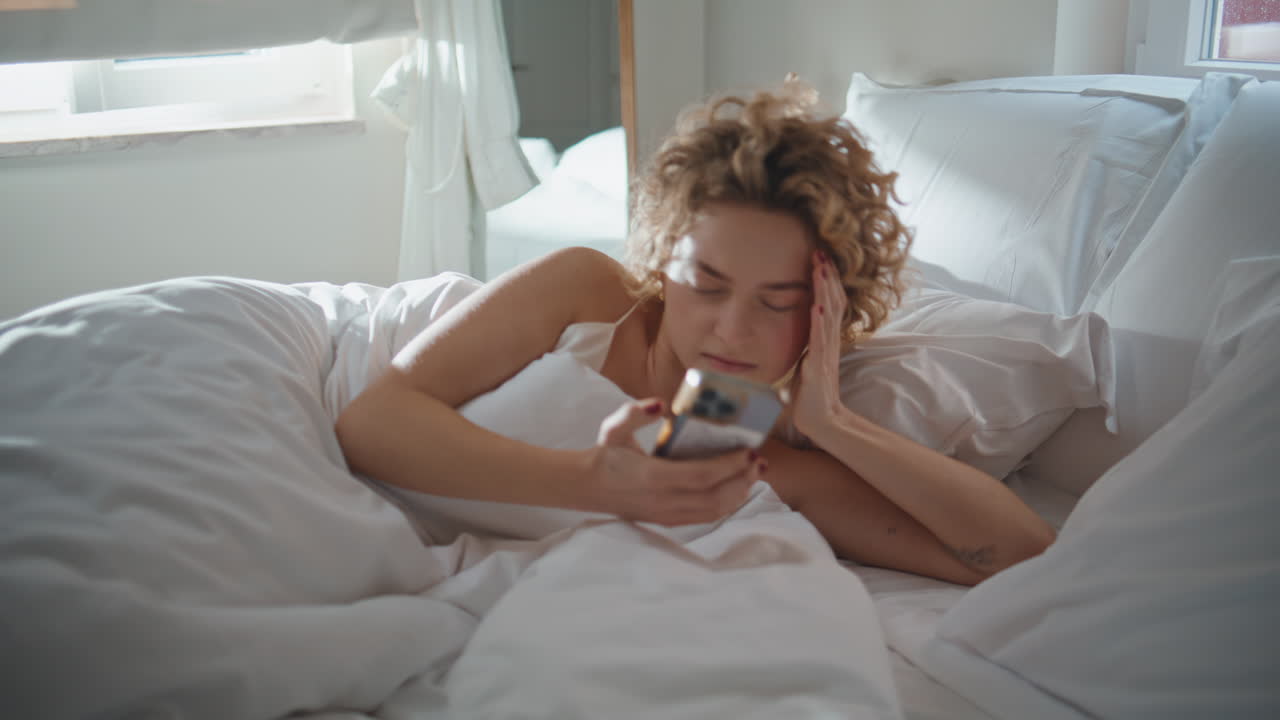 Woman checking cellphone bed in morning light. Serene girl waking up reading