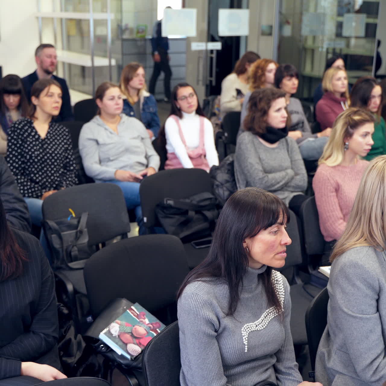 Young auditory listening to educator. Woman speaker holding a doll and telling the lecture to listeners in the modern hall.