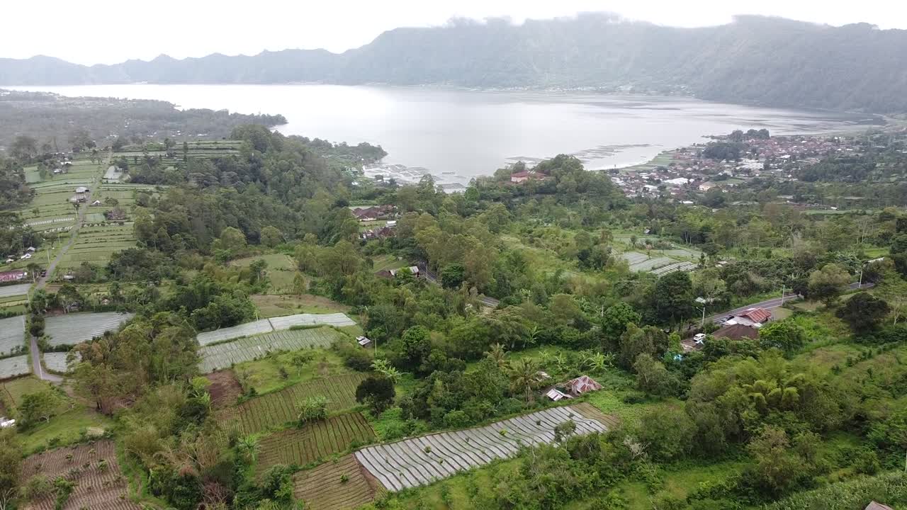 Drone footage showing a beautiful aerial view of green farmland and a calm lake surrounded by hills. Peaceful outdoor scenery on a cloudy day