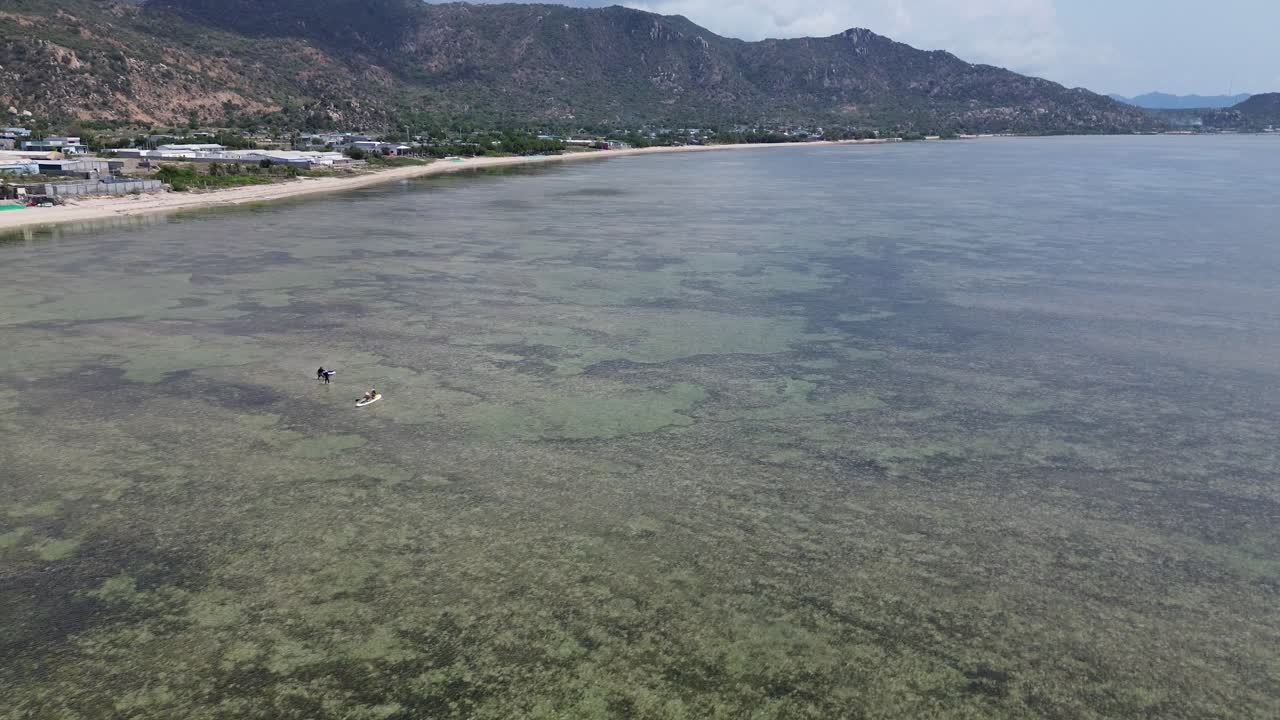 Drone flyr over padlebrettkjørere på stranden i Phan Rang, Vietnam, og panorerer deretter nedover for å avsløre det klare vannet, strandlinjen og strandaktiviteten