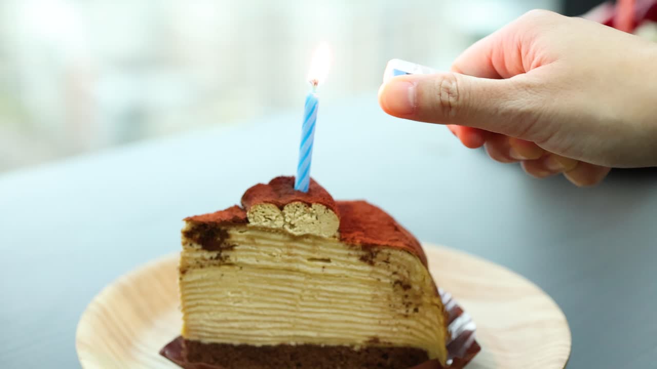 Hand lights blue candle on tiramisu crepe cake slice in bright, natural indoor setting