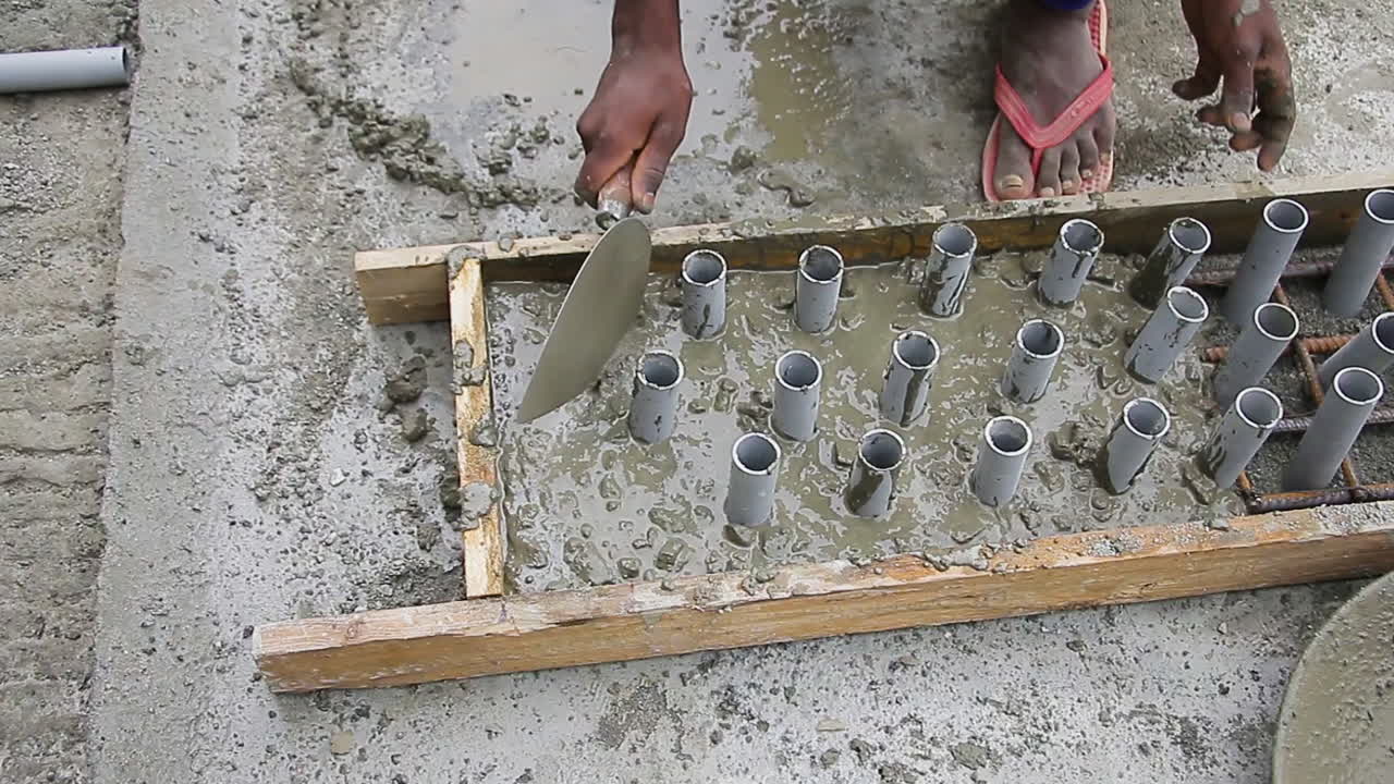 Indian mason worker placing and leveling wet concrete mix in slab around pipes in construction project