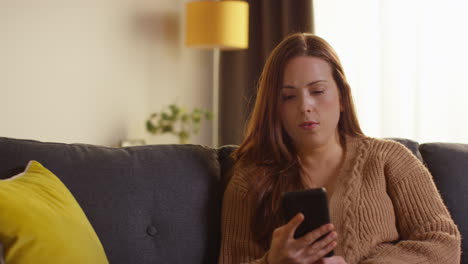 Puzzled-Woman-Sitting-On-Sofa-At-Home-Answering-Cold-Or-Nuisance-Call-On-Mobile-Phone-That-She-Doesn't-Recognise