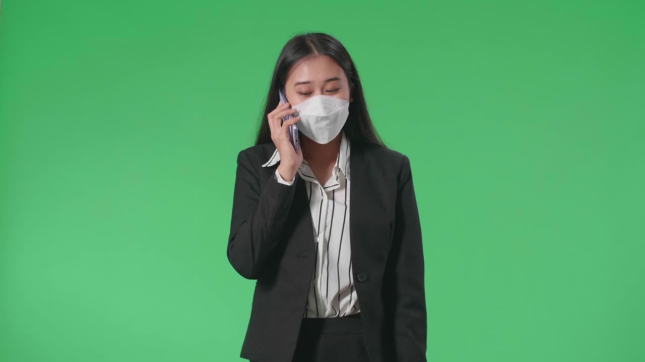 An Asian Business Woman Wearing Mask Talking On Mobile Phone While Walking In Front Of Green Screen In The Studio