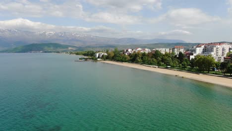 Drone-view-in-Albania-flying-over-Ohrid-blue-crystal-lake-in-Pogradec-beach-town-with-apartment-buildings-and-sandy-shore