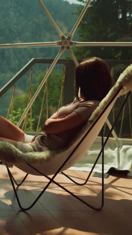 pensive woman enjoys incredible view of lake and mountains sitting in armchair with fur on glass balcony letting in suns rays. tropical holiday in modern tourist building