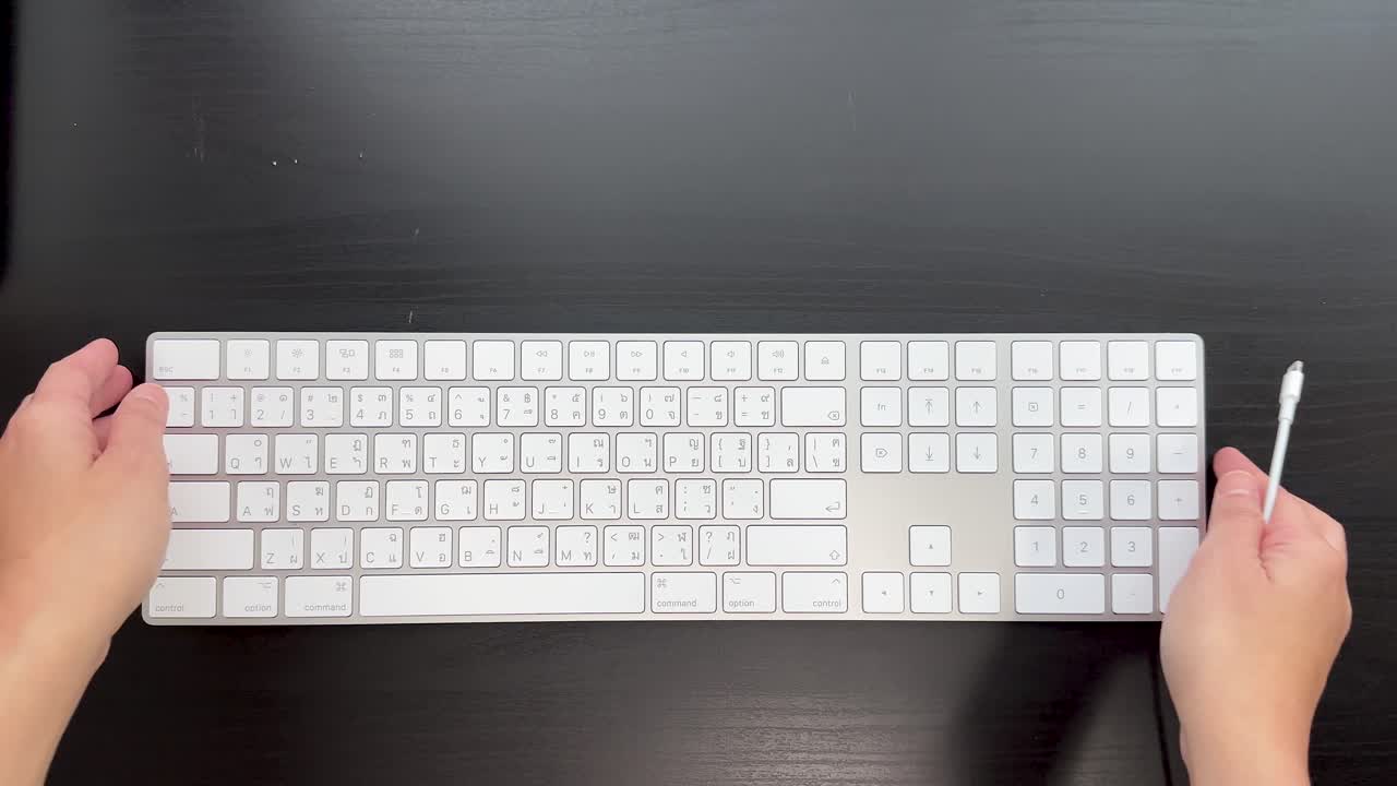 Hands plug cable into slim numeric keyboard on dark desk, bright even lighting, overhead perspective