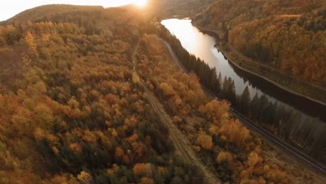 cinematic aerial diving car commercial establishing shot of cars driving on a road following a scenic water dam surrounded by lush autumn trees