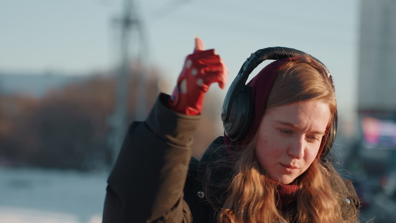 Femme blanche dans une rue enneigée, casque audio, pensive, ajustant son casque tout en écoutant de la musique, gants rouges et doudoune noire, longs cheveux bouclés captant le coucher de soleil doré, circulation urbaine et grand immeuble.