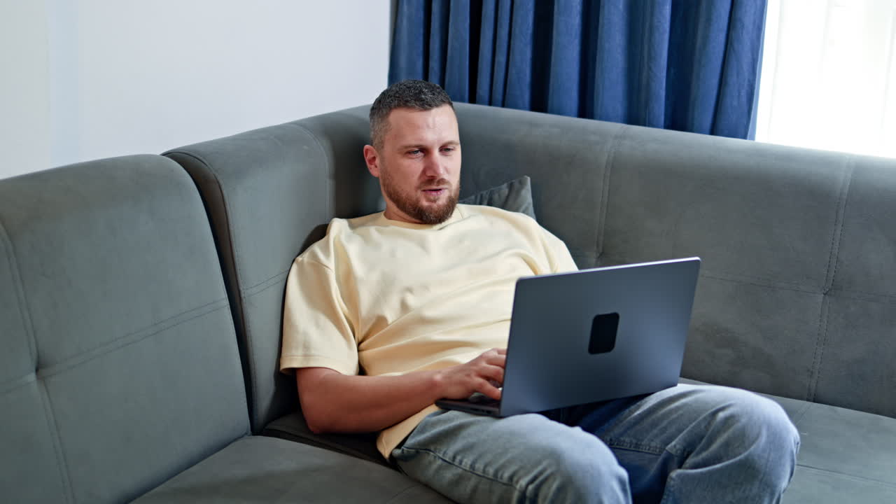 En mand arbejder på sin bærbare computer, siddende på sofaen i stuen i en moderne lejlighed med blå gardiner om dagen.
