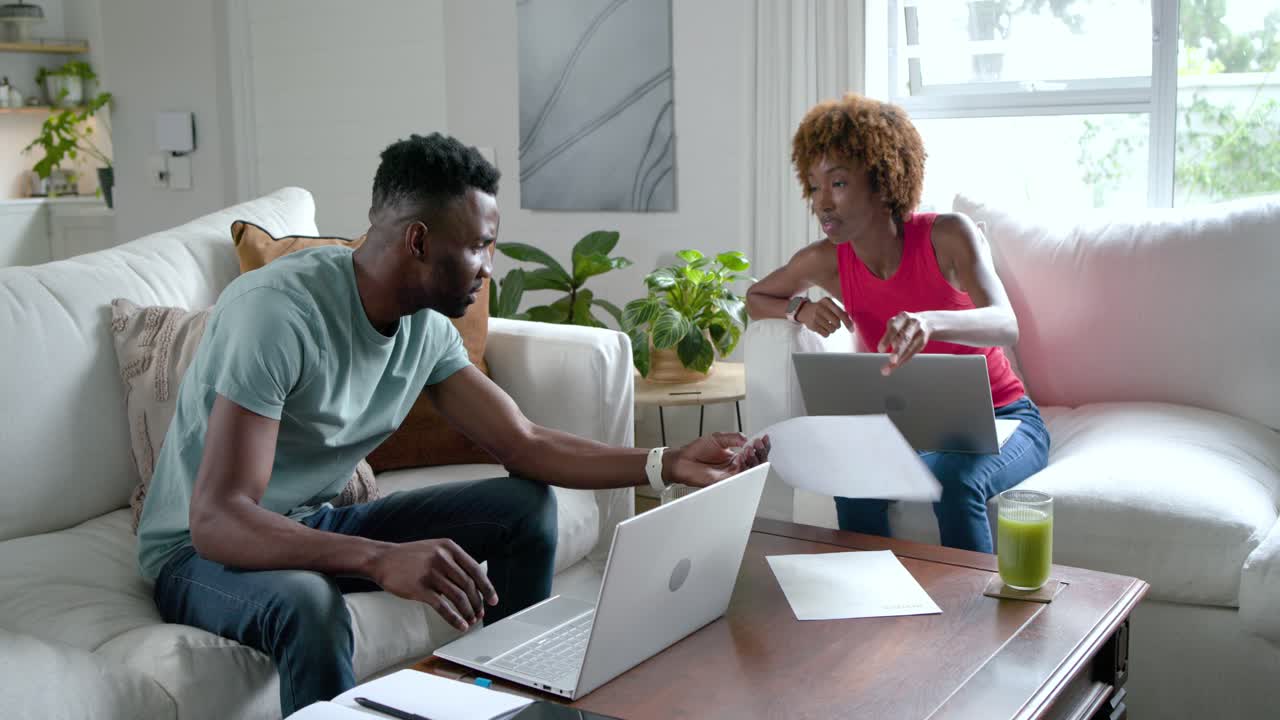 Couple afro-américain travaillant sur des ordinateurs portables à la maison, la femme feuilletant des pages, l'homme inspectant et triant